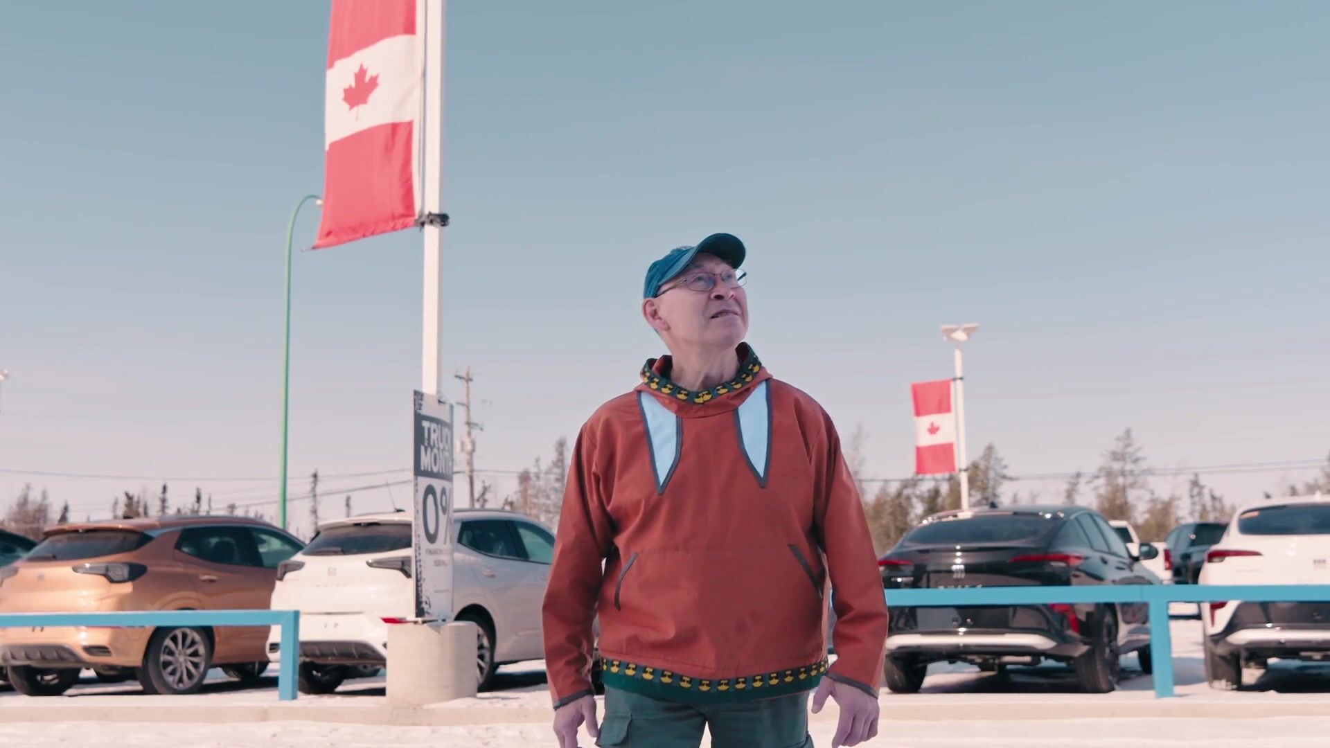 A man in a red sweater stands on the Yellowknife Motors lot in winter, a Canadian flag and rows of trucks behind him.