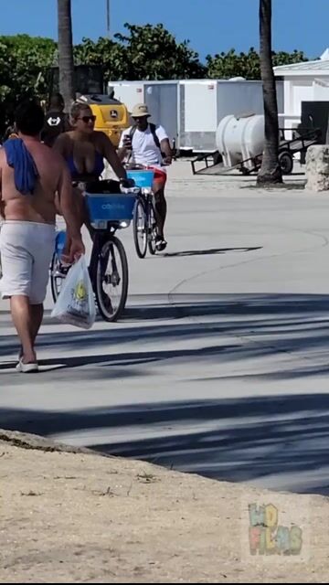 Woman in bikini top + shorts riding bike, covered, beach context